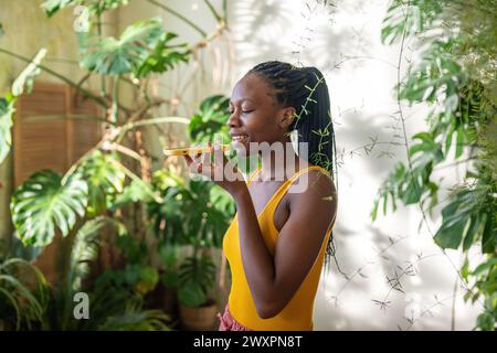 Pleased black young woman smiling records voice message on smartphone stands in tropical green room. Stock Photo