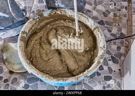 Construction worker tiler mixes cement adhesive mortar in bucket using paddle drill attachment. Stock Photo