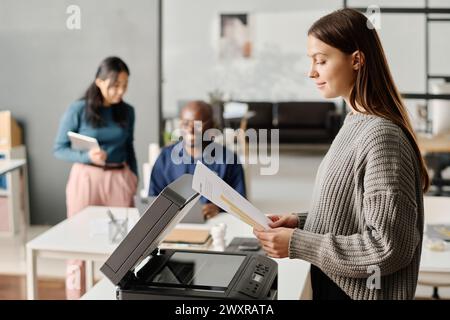 Side view of young Cacuasian manager wearing grey sweater standing in front of printer making copy of document, copy space Stock Photo