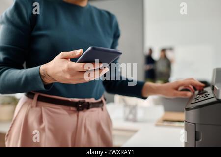Crop shot of unrecognizable woman wearing smart casual clothes holding smartphone using printer in modern office, dopy space Stock Photo