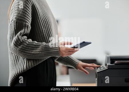 Side view crop shot of unrecognizable woman wearing gray sweater using smartphone while printing documents in office Stock Photo