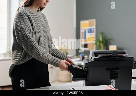 Crop shot of young Caucasian female office worker wearing gray sweater changing ink cartridge in printer, copy space Stock Photo