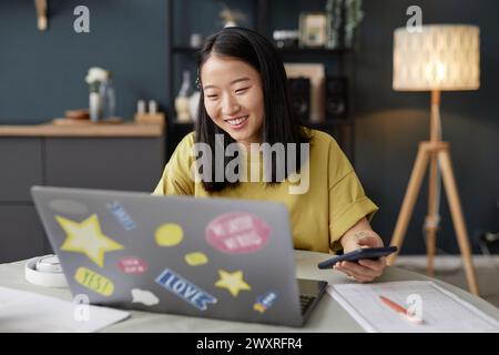 Joyful young adult Asian woman sitting at table in modern kitchen surfing Internet on laptop and smiling Stock Photo