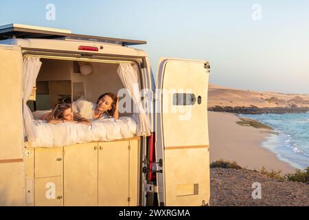 A couple of women are seen laid back on the bed of a trailer, gazing at the rising sun with a serene expression on their faces. Stock Photo