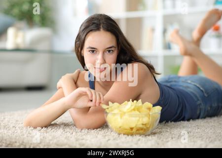 Beautiful woman looks at you eating potato chips lying on a carpet at home Stock Photo