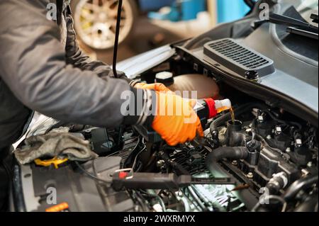 From above of crop anonymous male technician in uniform and gloves while standing in front of car with opened hood and pouring oil into engine in dayl Stock Photo