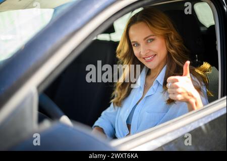 Smiling woman showing approval with a thumbs-up gesture while sitting in the driver's seat of a car Stock Photo