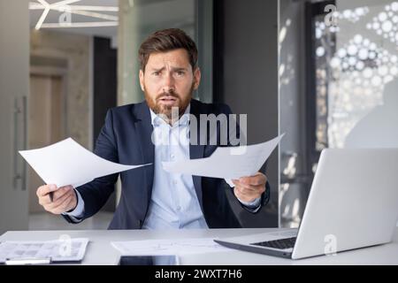 Confused businessman in a suit looks angrily at paperwork in a modern office setting, feeling frustrated Stock Photo