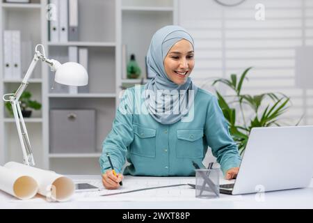 Smiling Muslim female architect working with blueprints and a laptop in a well-lit office space, conveying focus and professionalism. Stock Photo