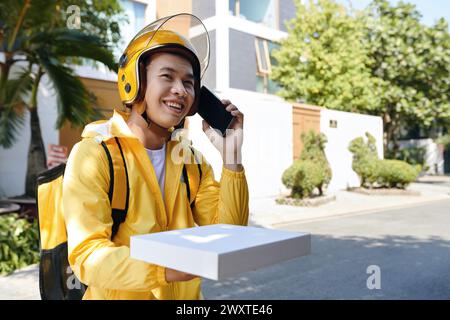 Smiling delivery man with box of pizza standing in font of customer house and making phone call Stock Photo