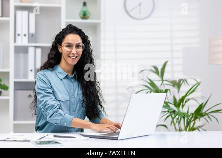 Cheerful Hispanic woman in a home office setting, smiling at the camera while working on her laptop with plants and a clock in the background. Stock Photo