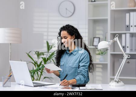 A cheerful Hispanic woman manages paperwork in her modern home office, exuding professionalism and positivity. Stock Photo