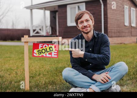 Portrait of smiling young man pointing to sold sign outside house. Happy caucasian man buying new home. Stock Photo