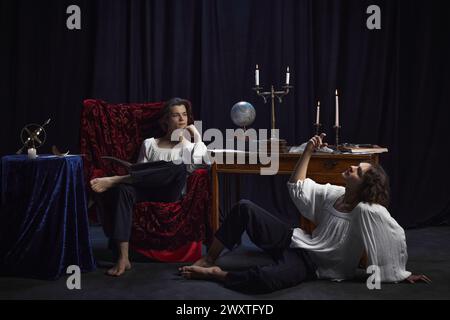 Creative portrait of two medieval persons sitting in room with vintage furniture and drinking wine while writing novel. Stock Photo