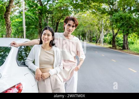 Asian couple standing next to their car on a roadside with lush greenery Stock Photo