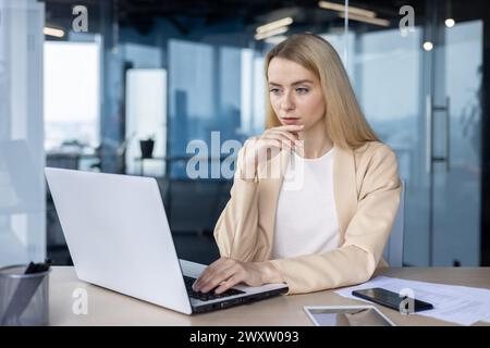 Focused businesswoman contemplating while working on laptop in modern office setting, representing serious thinking. Stock Photo