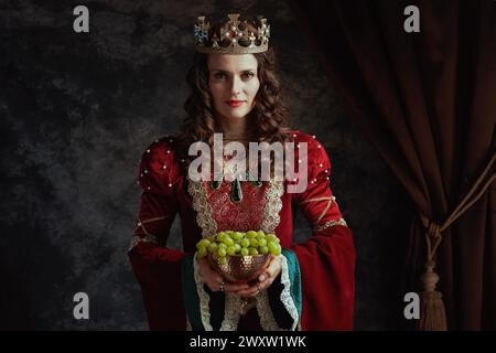 smiling medieval queen in red dress with plate of grapes and crown on dark gray background. Stock Photo