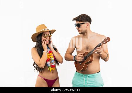 Multicultural couple enjoying summer vibes at the beach. Latin woman in bikini looks surprised as man gazes at her while holding a ukulele on white ba Stock Photo