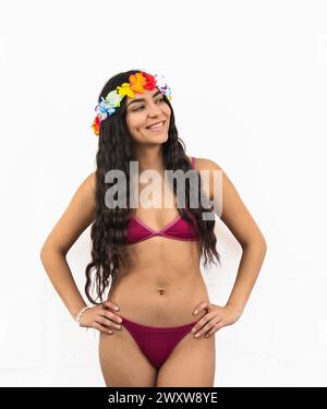 Cheerful young Latina woman poses confidently in a bikini with hands on hips and a flower crown, radiating joy and enjoying the summer vibes on white Stock Photo