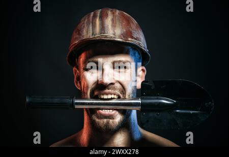 Heavy industry and mining. Male industrial worker, miner in safety hard hat with shovel in teeth. Closeup portrait of serious professional builder in Stock Photo