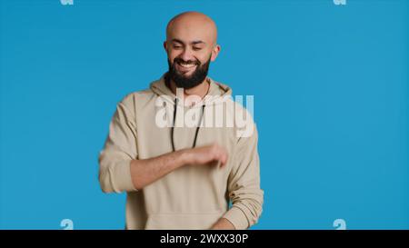 Middle eastern adult showing groovy dance moves in studio, having fun feeling funky and relaxed over blue background. Young guy in studio dancing and feeling confident, cool man. Camera 1. Stock Photo
