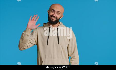 Middle eastern guy greeting people in front of camera, waving hello and smiling over blue background. Young person posing with confidence in studio, feeling confident and joyful. Camera 1. Stock Photo