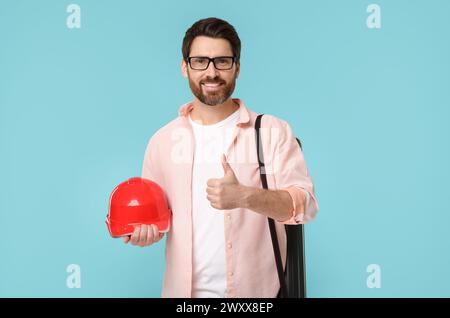 Architect with drawing tube and hard hat showing thumbs up on light blue background Stock Photo