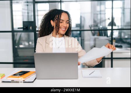 Working with documents. Positive brazilian or hispanic business woman, company employee, financial ceo, sits at a work desk in modern office, working in a laptop, studying financial documents, smile Stock Photo