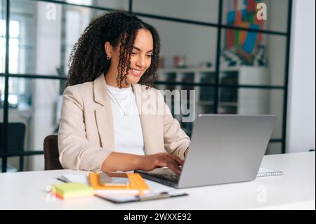 Working in the office. Positive business lady working on laptop in office, brazilian businesswoman in formal jacket sitting at a work desk in a modern space, working on a project in a laptop, smiling Stock Photo