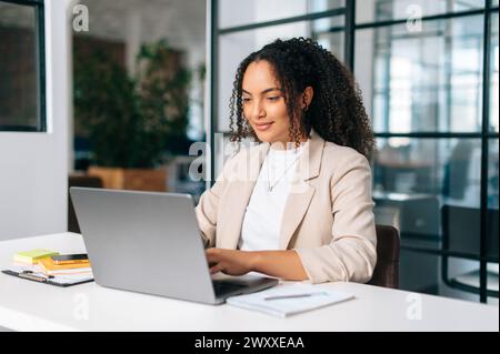 Woman uses laptop. Successful curly haired hispanic or brazilian woman in stylish elegant clothes, office employee, secretary or hr manager, using a laptop while sitting at her workplace, working Stock Photo