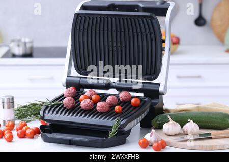 Electric grill with meatballs, tomatoes and vegetables on white table in kitchen Stock Photo