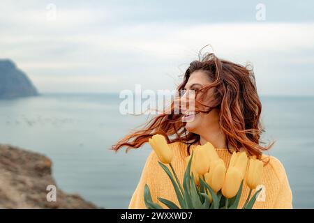 Portrait of a happy woman with hair flying in the wind against the backdrop of mountains and sea. Holding a bouquet of yellow tulips in her hands Stock Photo