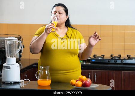 Overweight fat indian woman drinking a fruit juice in a kitchen. Jar and fruits placed on table.healthy eating concept. Copy Space Stock Photo