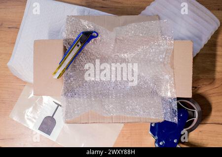 Open box with wrapped items, adhesive tape, scissors, paper and bubble wrap on wooden table, flat lay. Stock Photo
