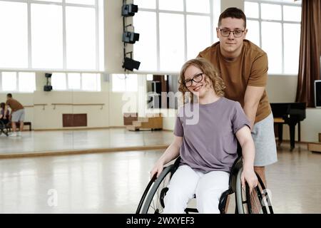 Medium long portarit of cheerful young woman in wheelchair and young man standing behind her in dance studio, copy space Stock Photo