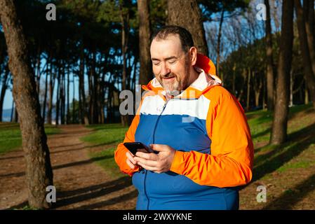 A middle-aged man engaging with his smartphone, dressed in a colorful sports jacket, stands in a park with tall trees in the background Stock Photo