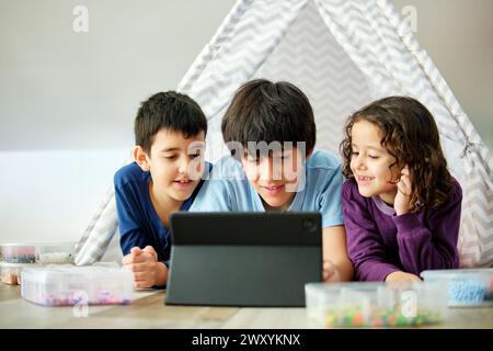 Two boys and a girl engrossed in a digital tablet, with colorful hama beads nearby, inside a cozy play tent. Stock Photo