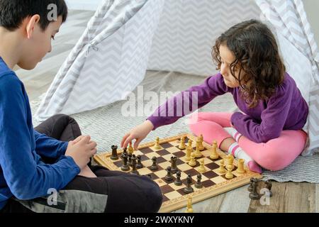 Two children, a boy and a girl, are carefully contemplating their next move while playing chess on a wooden board sitting on the floor Stock Photo