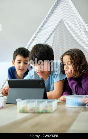 Two boys and a girl, siblings, explore content on a digital tablet together, with focus and curiosity, in a cozy home playroom setting. They are looki Stock Photo