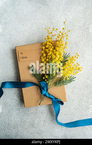Top view of charming kraft paper gift box tied with a blue satin ribbon, complemented by vibrant mimosa flowers, set against a textured grey backgroun Stock Photo