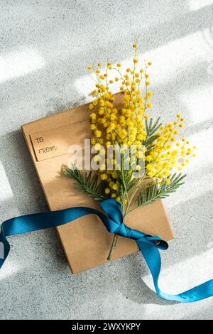 Top view of brown paper-wrapped gift box tied with a vibrant blue ribbon, featuring a bouquet of yellow mimosa flowers, placed on a textured gray back Stock Photo