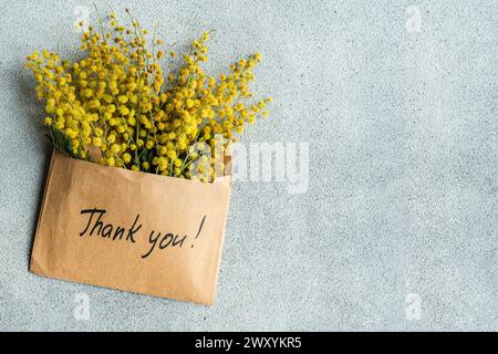 Top view of kraft paper envelope with Thank you! written on it, overflowing with bright yellow mimosa flowers on a grey background. Stock Photo