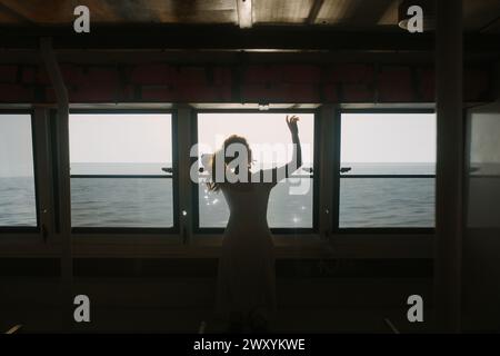 A woman stands in silhouette inside a ferry, reaching toward the sunlight streaming in through the windows with the ocean in the background Stock Photo