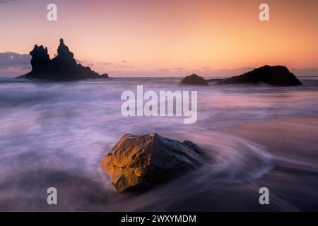 The setting sun casts a golden glow over Playa de Benijo, highlighting the sea's rhythmic dance around the iconic rock formations Stock Photo