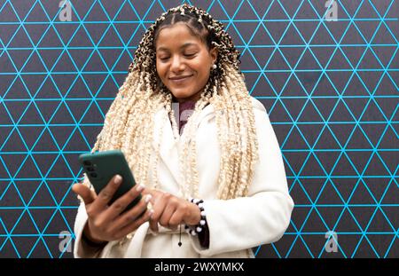 A content Black woman engrossed in her mobile phone, with a joyful expression, stands against a geometric backdrop in the city. Stock Photo