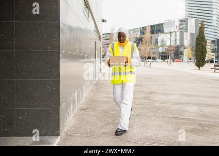 A black woman wearing a hijab and safety vest walks with a package in an urban setting. Stock Photo