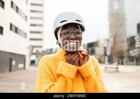 A joyful Black woman wearing a safety helmet and glasses, smiles broadly in the city. Stock Photo