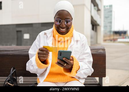 A joyful Black woman in a hijab and glasses sits outdoors, typing on her phone with a credit card in her hand. Stock Photo