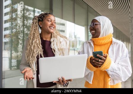 Two African American women share a cheerful moment, one holding a laptop, the other using a mobile phone, engaging in a lively conversation on a city Stock Photo