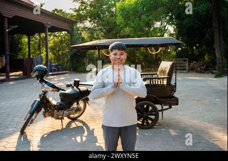 Smiling young man performing a traditional greeting with hands pressed together, standing outdoors with a motorcycle and tuk-tuk in the background Stock Photo